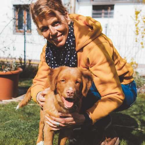 Eine Person in einem Garten, die mit einem braunen Hund lächelt.