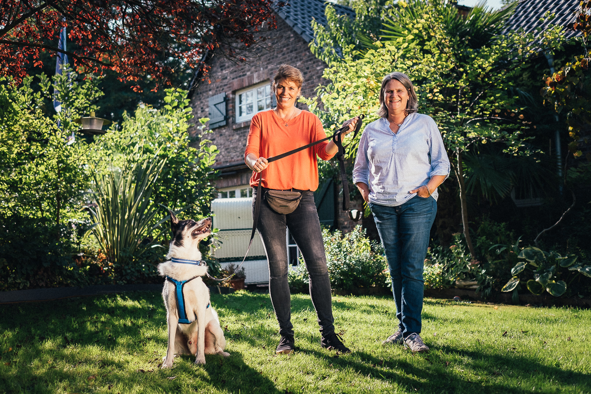 Zwei Menschen stehen mit einem Hund auf einem grasbewachsenen Garten.