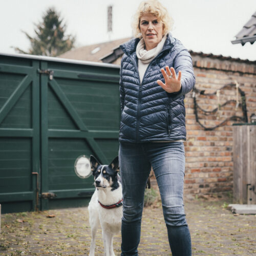 Frau in blauer Jacke, Hand erhoben, steht neben einem schwarz-weißen Hund im Hof.