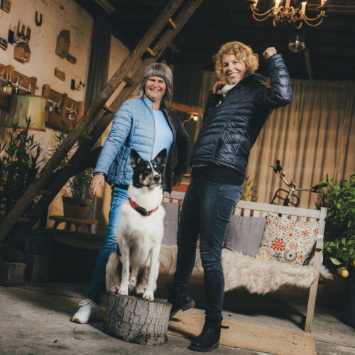 Zwei Personen in Winterjacken posieren drinnen, ein Hund sitzt auf einem Baumstamm. Wohnzimmeratmosphäre.