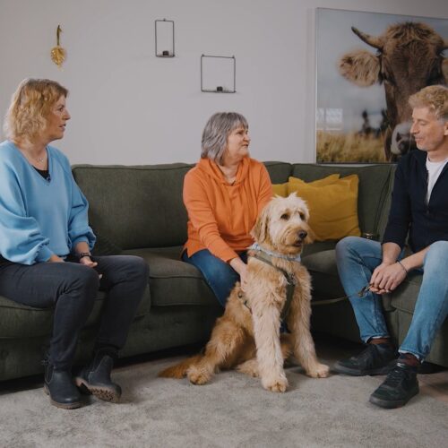 Drei Personen auf einem Sofa, mit einem Hund in der Mitte. Wohnzimmer mit Fenster und Pflanze.