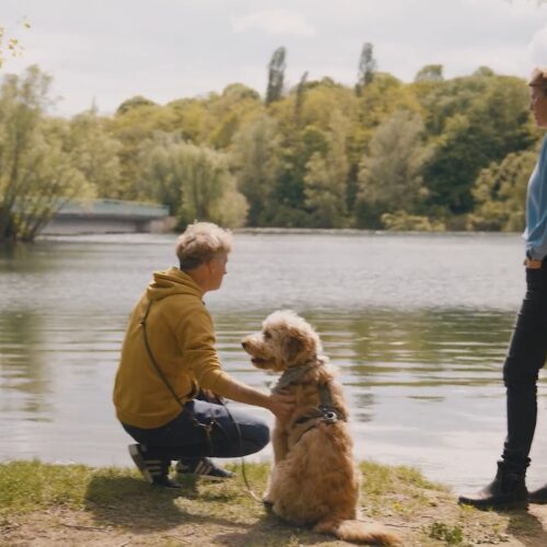 Zwei Personen mit Hund am Ufer eines Sees, umgeben von Bäumen.